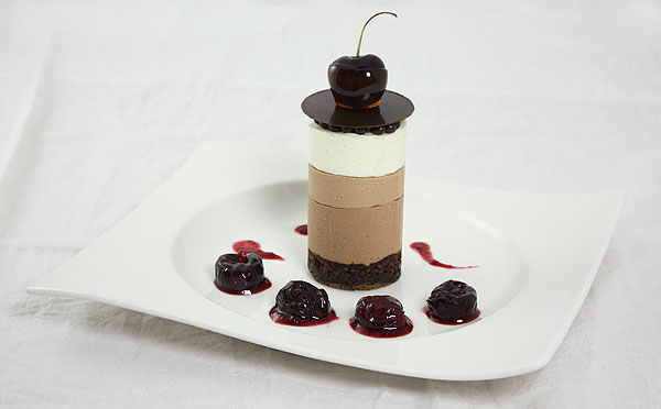 Mini Chocolate Mousse decorated with crispearls, chocolate disc and caramel dipped cherry on white dessert dish decorated with cherry purée and poached cherries.