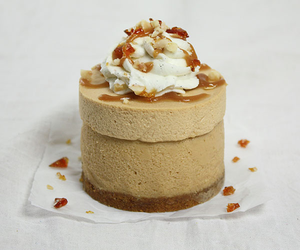 Close up photo of a Mini Dulce de Leche Cheesecake topped with swirl of whipped cream drizzled with caramel and sprinkled with Hazelnut Praline displayed on parchment square.