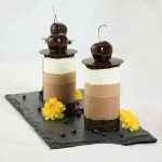Mini Triple Chocolate Mousse Cakes decorated with Crispearls, Chocolate Disc, Caramel Dipped Cherries and Edible Flowers displayed on black slate board.
