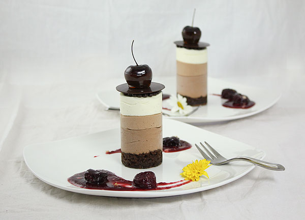 Two plated Mini Triple Chocolate Mousse Cakes on white dessert dishes that have been decorated with cherry purée and poached cherries.
