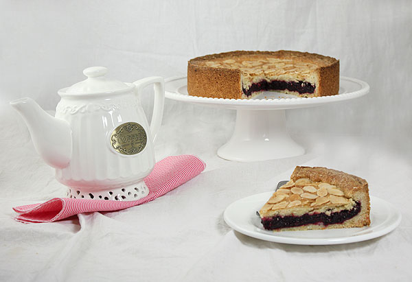 Photo of Blackberry Shortbread Cake with slice removed displayed on white cake stand with slice displayed on white dessert dish set in front with white coffee pot set to one side.