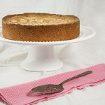Photo of Blackberry and Almond Shortbread Cake displayed on white ruffled edge cake stand with pink and white striped cloth napkin and antique cake server.