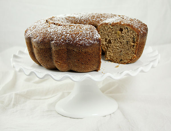Pear and Walnut Quick Bread dusted with confectioners' sugar with slice removed displayed on white cake stand.